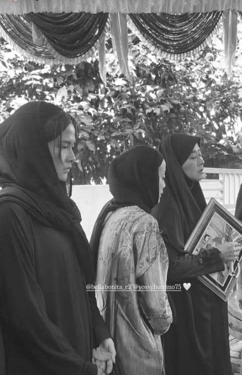 Beberapa perempuan berpakaian hitam dan berjilbab berdiri khidmat di pemakaman, salah satunya memegang bingkai foto almarhum.
