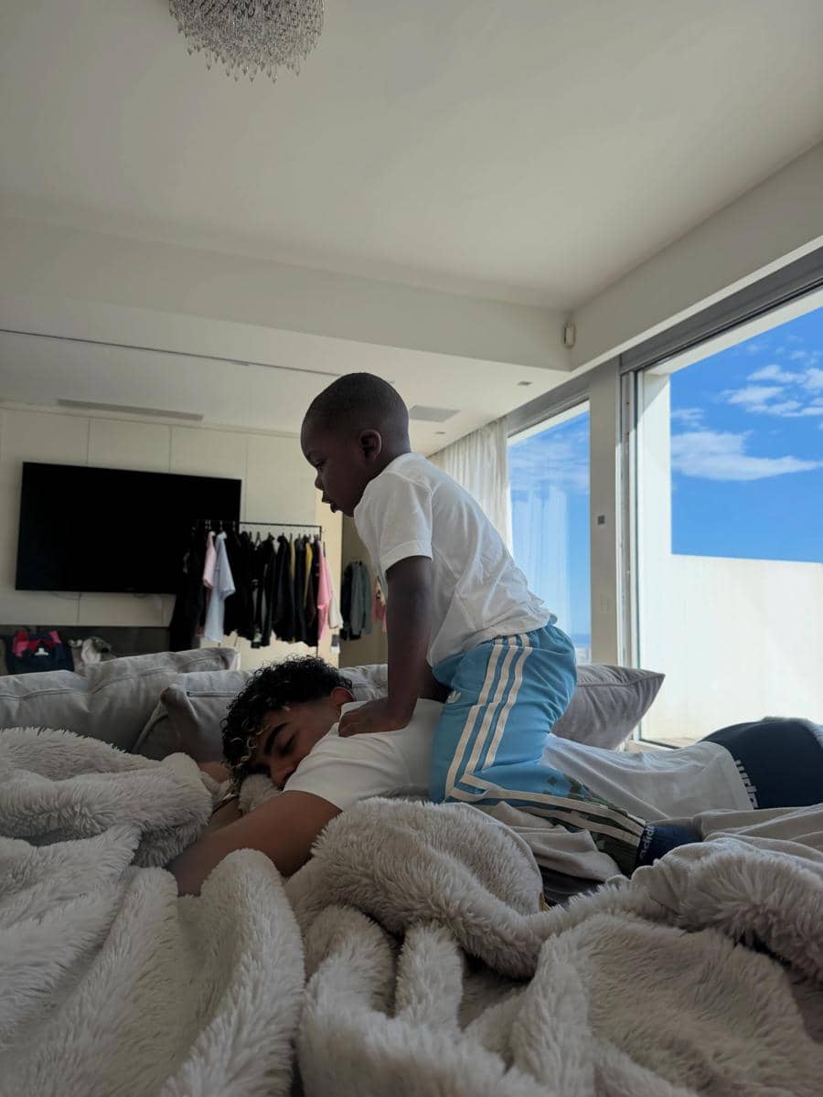Seorang anak kecil duduk di punggung seorang remaja yang sedang berbaring di sofa, di dalam ruangan dengan jendela besar dan langit cerah di luar.