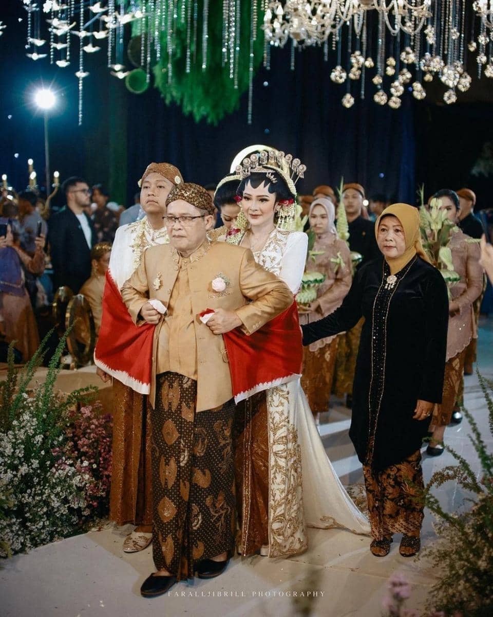 Seorang pengantin wanita mengenakan busana adat Jawa berjalan bersama ayahnya di acara pernikahan dengan dekorasi mewah dan tradisional.