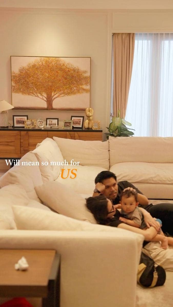Sebuah keluarga sedang bersantai di ruang tamu rumah baru dengan sofa besar berwarna krem dan lukisan pohon di dinding.