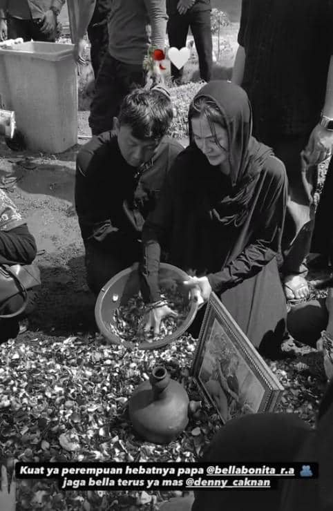 Seorang perempuan berpakaian hitam menabur bunga di atas makam sambil didampingi seorang pria dalam suasana pemakaman yang khidmat.
