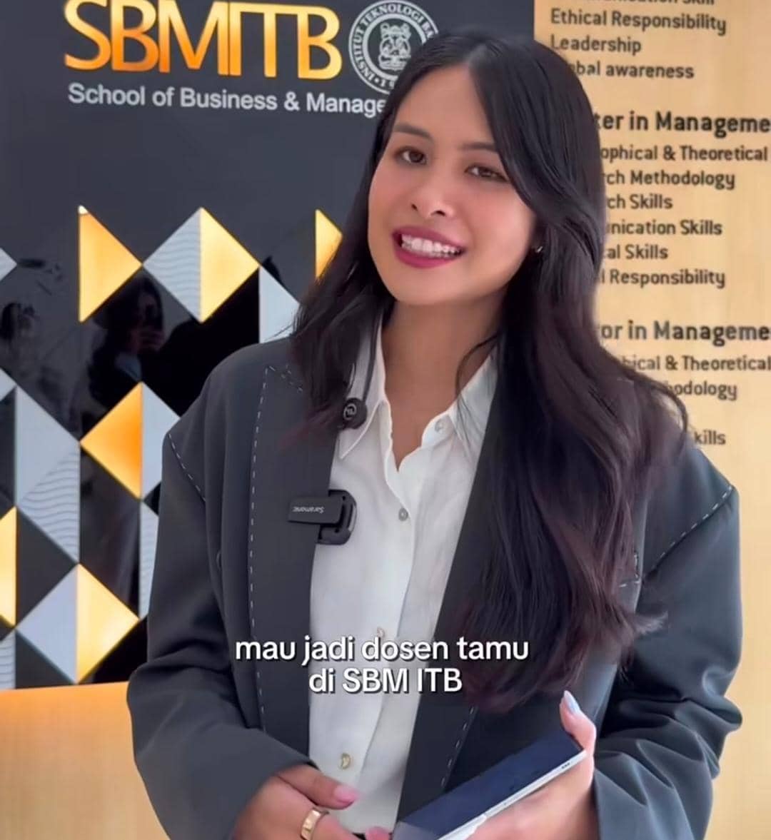 Seorang perempuan mengenakan jas abu-abu dan kemeja putih berdiri di depan latar bertuliskan SBM ITB, tersenyum sambil memegang ponsel.