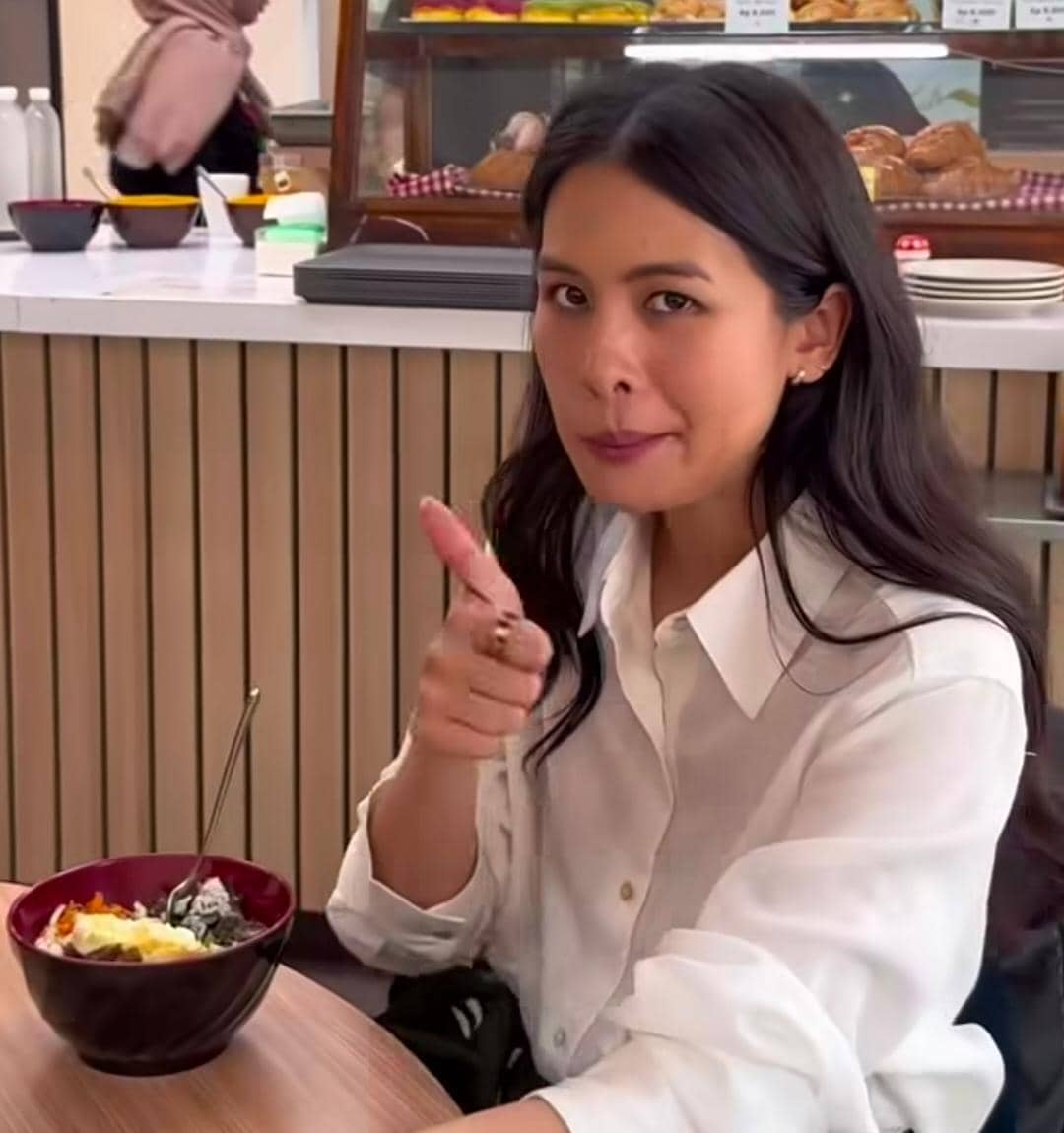 Seorang perempuan berambut panjang mengenakan kemeja putih duduk di meja kafe sambil menunjuk dengan ekspresi ceria di depan semangkuk makanan.