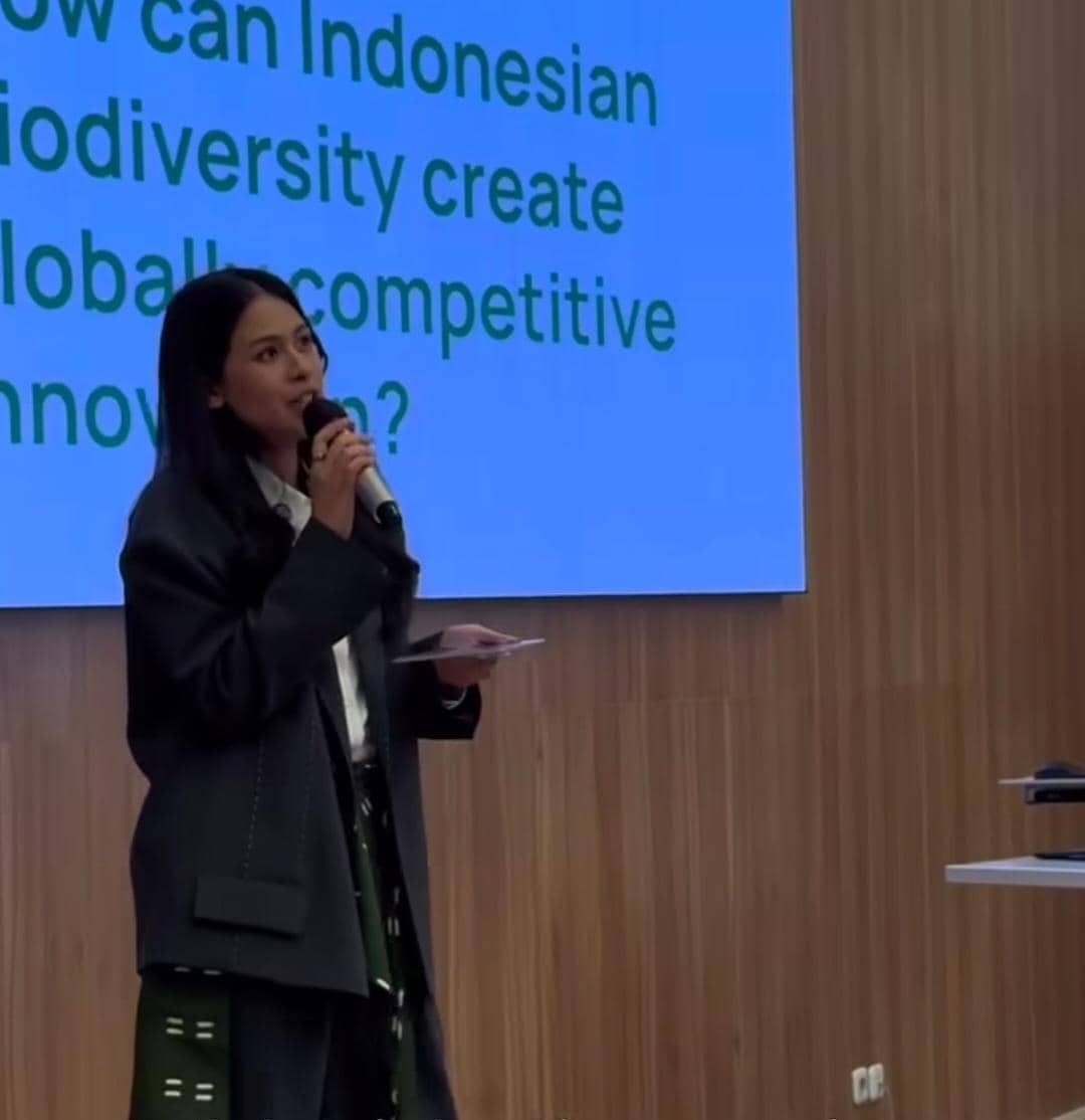 Seorang perempuan berbicara menggunakan mikrofon di depan layar presentasi bertuliskan topik tentang inovasi dan keanekaragaman hayati Indonesia.