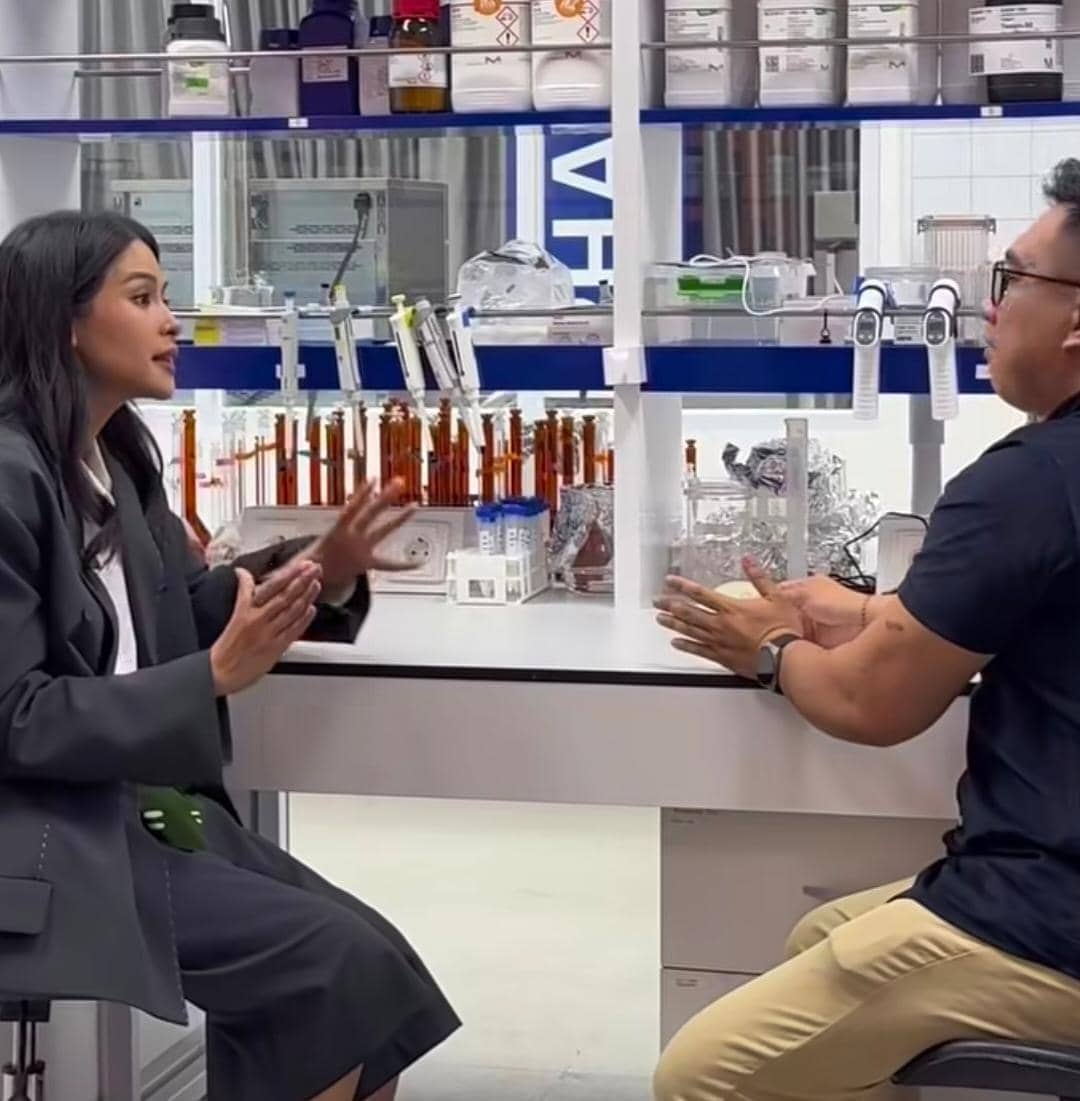 Seorang wanita berbicara dengan seorang pria di laboratorium dengan peralatan kimia di atas meja dan rak di belakang mereka.