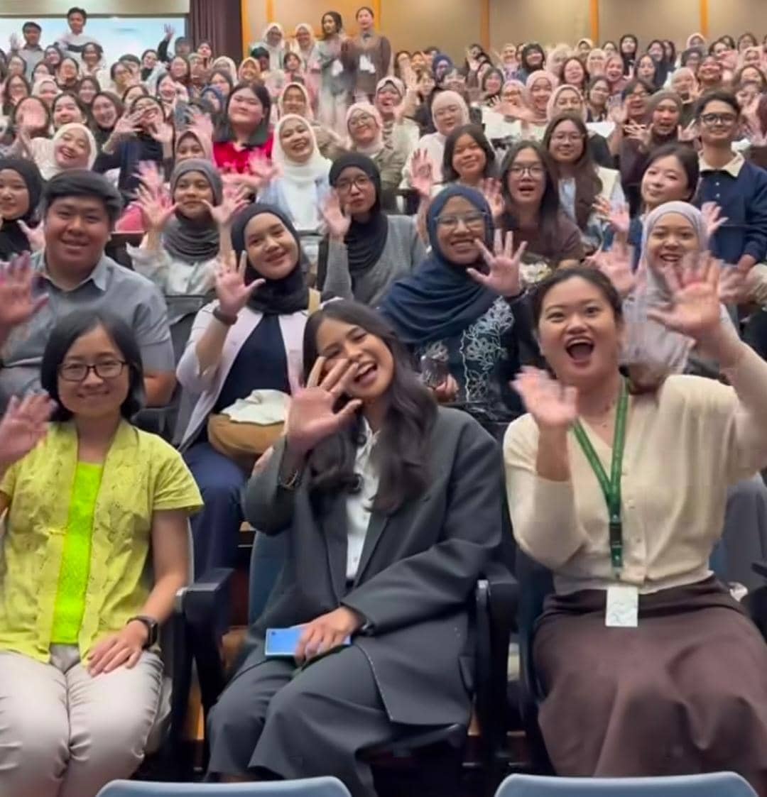 Seorang dosen tamu duduk di barisan depan bersama mahasiswa di ruang kuliah ITB, semuanya tersenyum dan melambaikan tangan ke kamera.