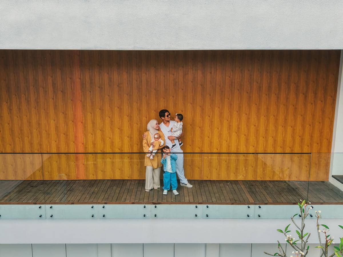 Sebuah keluarga berdiri di balkon vila modern dengan dinding kayu di Bali, menikmati waktu bersama dengan suasana hangat dan cerah.