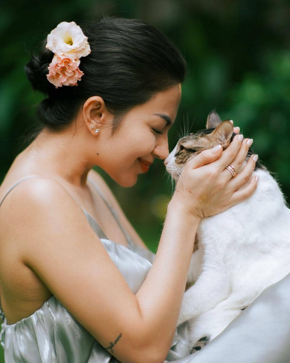 Seorang wanita bergaun satin abu-abu tersenyum sambil menyentuhkan hidungnya dengan seekor kucing putih belang cokelat di taman hijau.