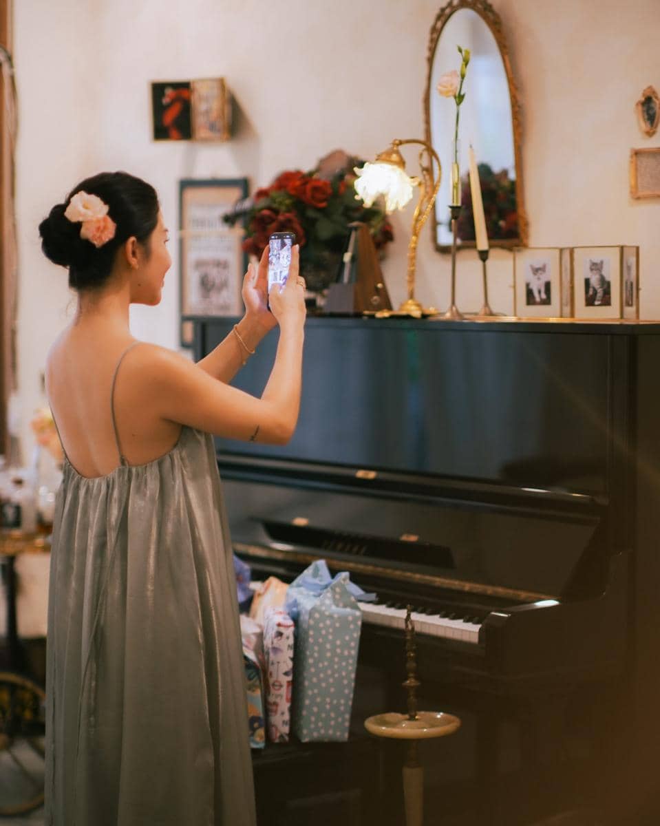 Seorang wanita bergaun abu-abu memotret dengan ponsel di depan piano hitam yang dihiasi bunga, lilin, dan bingkai foto kucing.
