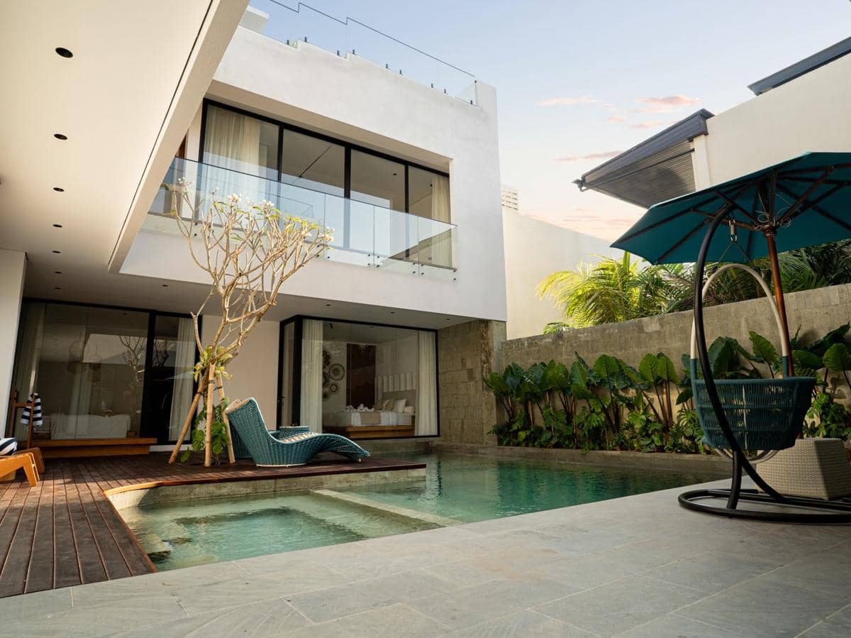 Vila modern dua lantai di Bali dengan kolam renang pribadi, kursi santai, dan area terbuka yang dikelilingi tanaman tropis.