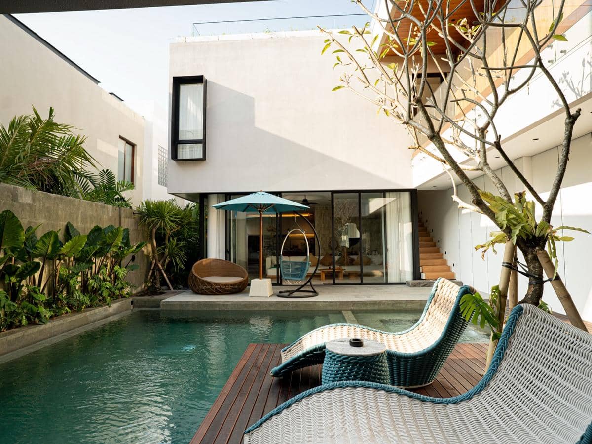Kolam renang pribadi dengan kursi santai rotan di vila modern dua lantai bergaya tropis yang dikelilingi tanaman hijau di Bali.