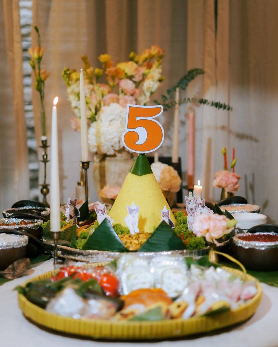 Tumpeng nasi kuning dengan hiasan angka lima dan gambar kucing di atas meja, dikelilingi lilin serta bunga berwarna lembut.