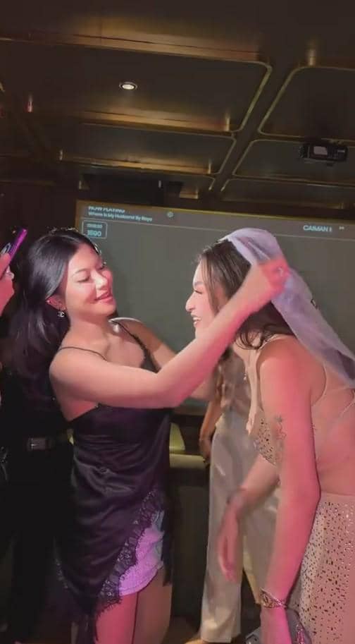 Seorang wanita mengenakan gaun hitam membantu memasangkan veil pengantin pada wanita lain yang tersenyum di acara bridal shower.