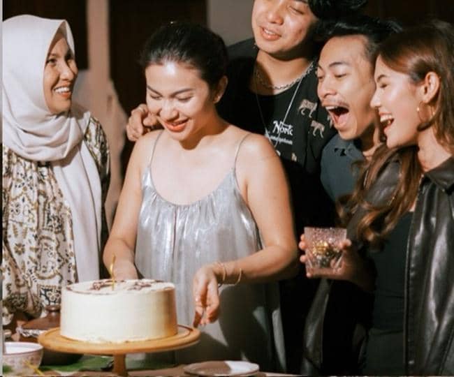 Seorang wanita mengenakan gaun perak tersenyum di depan kue ulang tahun bersama empat orang teman yang ikut merayakan dengan gembira.