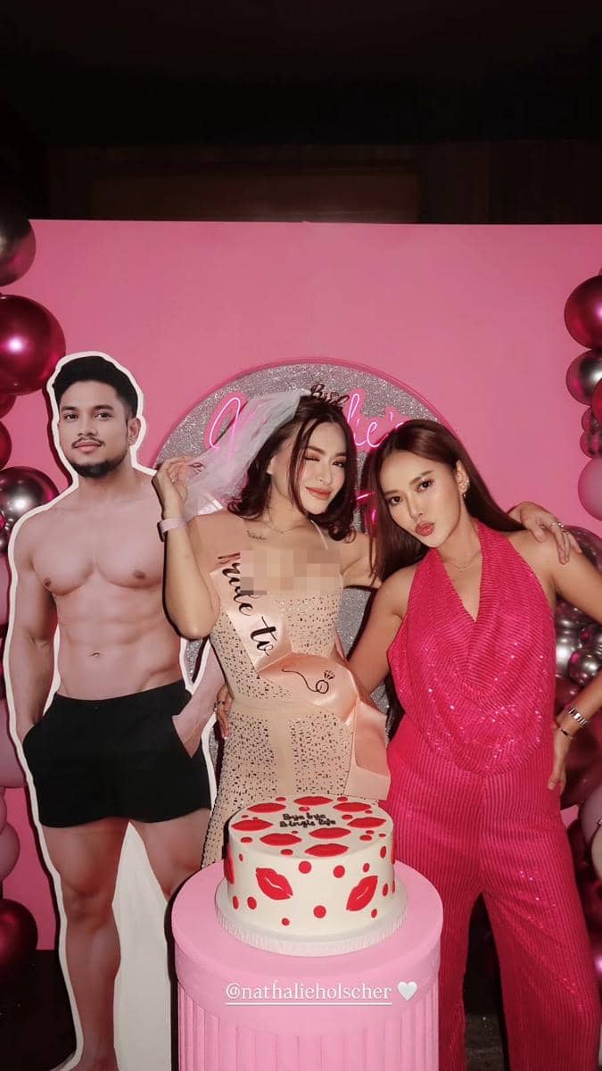 Dua wanita berpose di pesta bridal shower bertema merah muda dengan kue dekoratif dan latar balon serta poster pria bertelanjang dada.