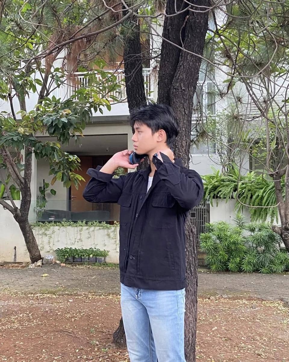 Seorang pria berdiri di luar ruangan mengenakan jaket hitam dan celana jeans biru, dengan latar pepohonan dan bangunan rumah.