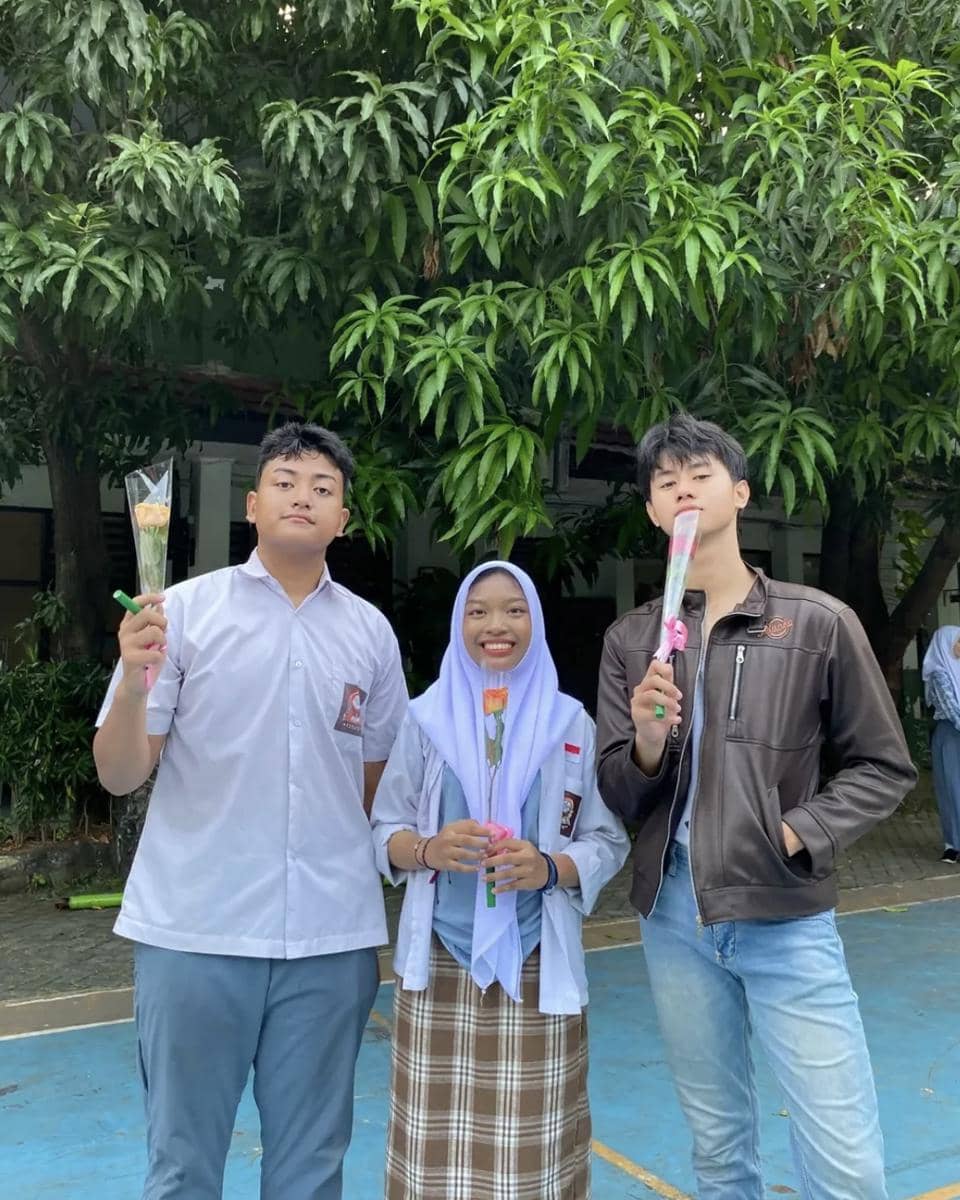 Tiga siswa berdiri di luar ruangan sambil memegang bunga mawar, dua mengenakan seragam sekolah dan satu memakai jaket kulit cokelat.