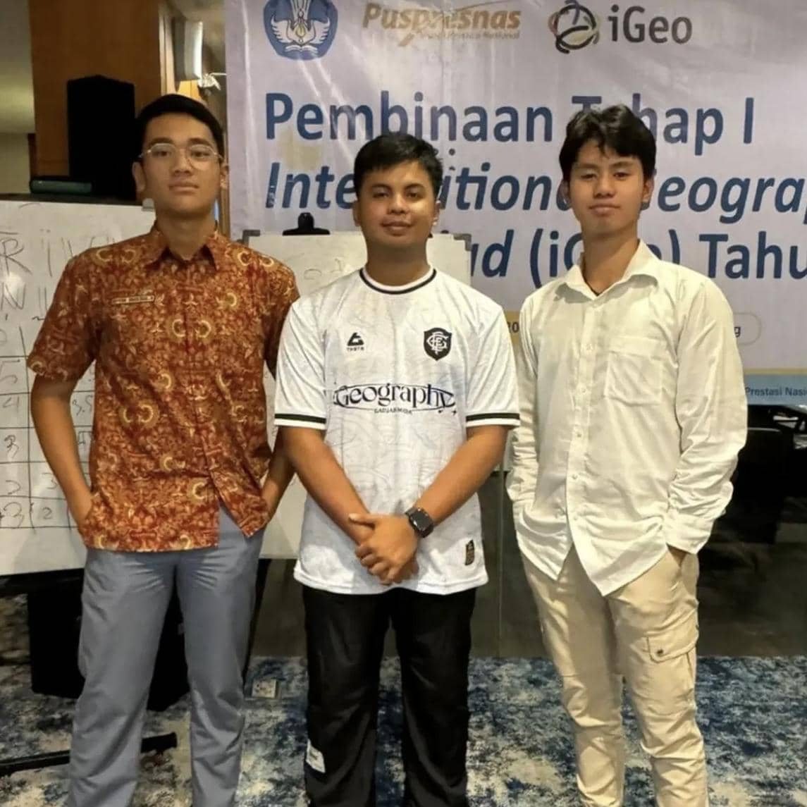 Tiga peserta muda berdiri bersama di depan spanduk bertuliskan Pembinaan Tahap I International Geography Olympiad di sebuah ruangan.