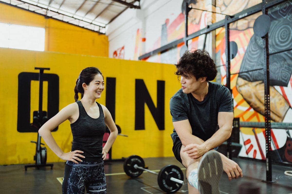 Dua orang berolahraga di gym dengan latar dinding kuning dan mural warna-warni, bersiap melakukan latihan kekuatan dan kebugaran.