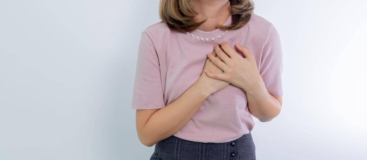 Seorang wanita mengenakan blus ungu muda memegangi dadanya dengan kedua tangan, menggambarkan gejala serangan jantung.