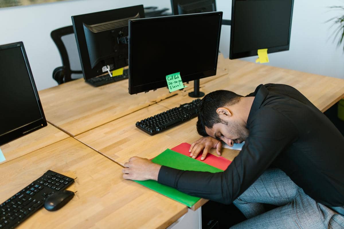 Seorang pria mengenakan kemeja hitam tertidur di meja kantor dengan beberapa monitor komputer dan map berwarna di depannya.