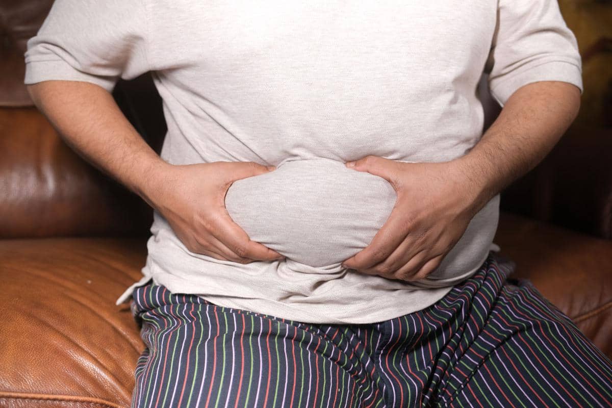 Seorang pria duduk di sofa sambil memegang perut buncitnya dengan kedua tangan, mengenakan kaus abu-abu dan celana bergaris.