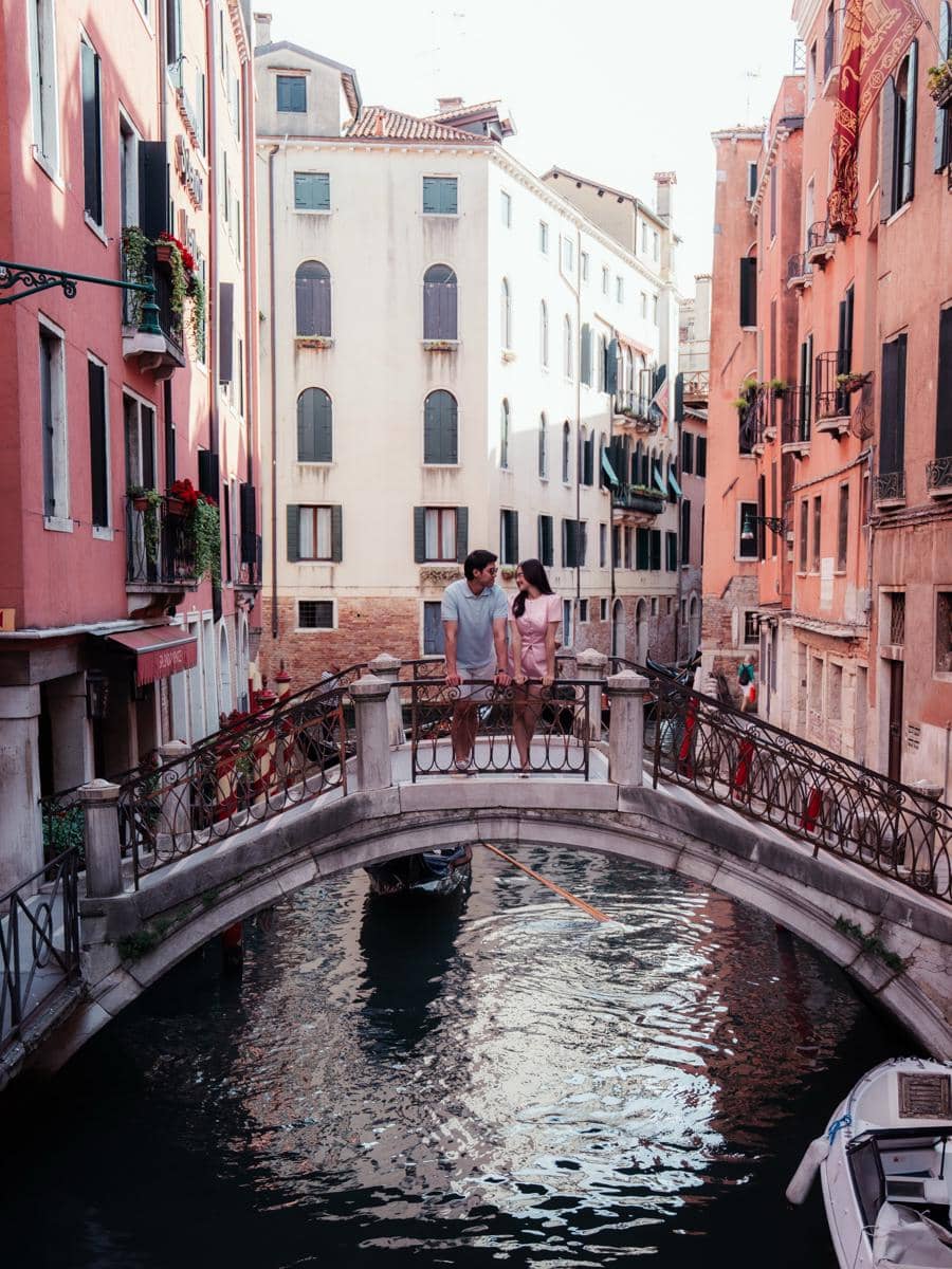 Sepasang kekasih berdiri di atas jembatan kecil di Venice dengan latar bangunan berwarna pastel dan kanal yang tenang.