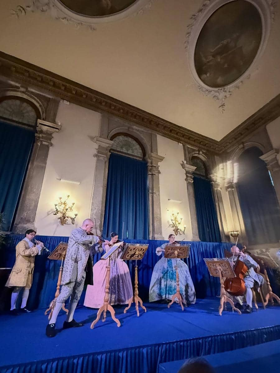 Sekelompok musisi memainkan alat musik klasik dengan kostum abad ke-18 di atas panggung dalam ruangan bergaya Eropa dengan tirai biru.