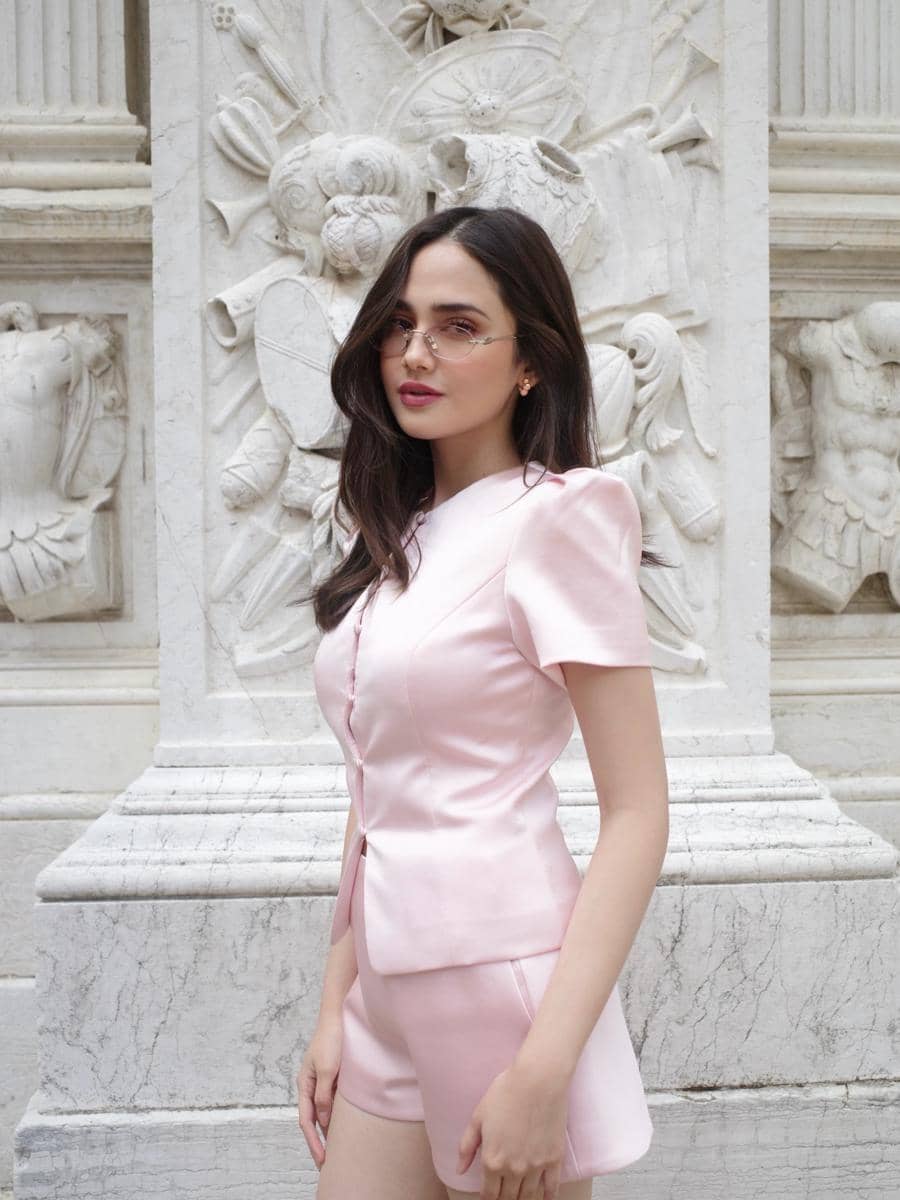 Seorang wanita mengenakan setelan berwarna pink muda dan kacamata kecil berpose di depan dinding marmer berukir di Venice.