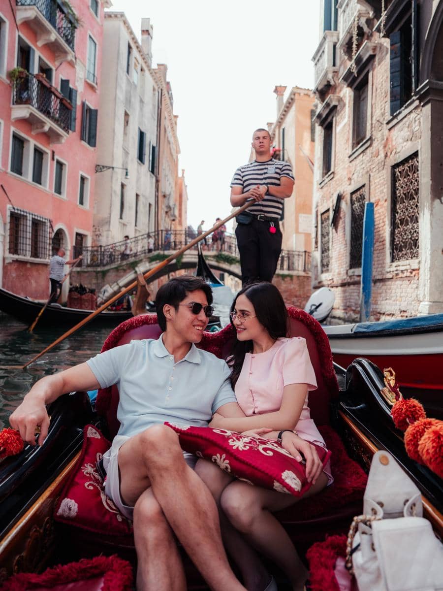 Sepasang kekasih duduk di gondola sambil tersenyum dan saling menatap di kanal Venesia dengan latar bangunan klasik Italia.