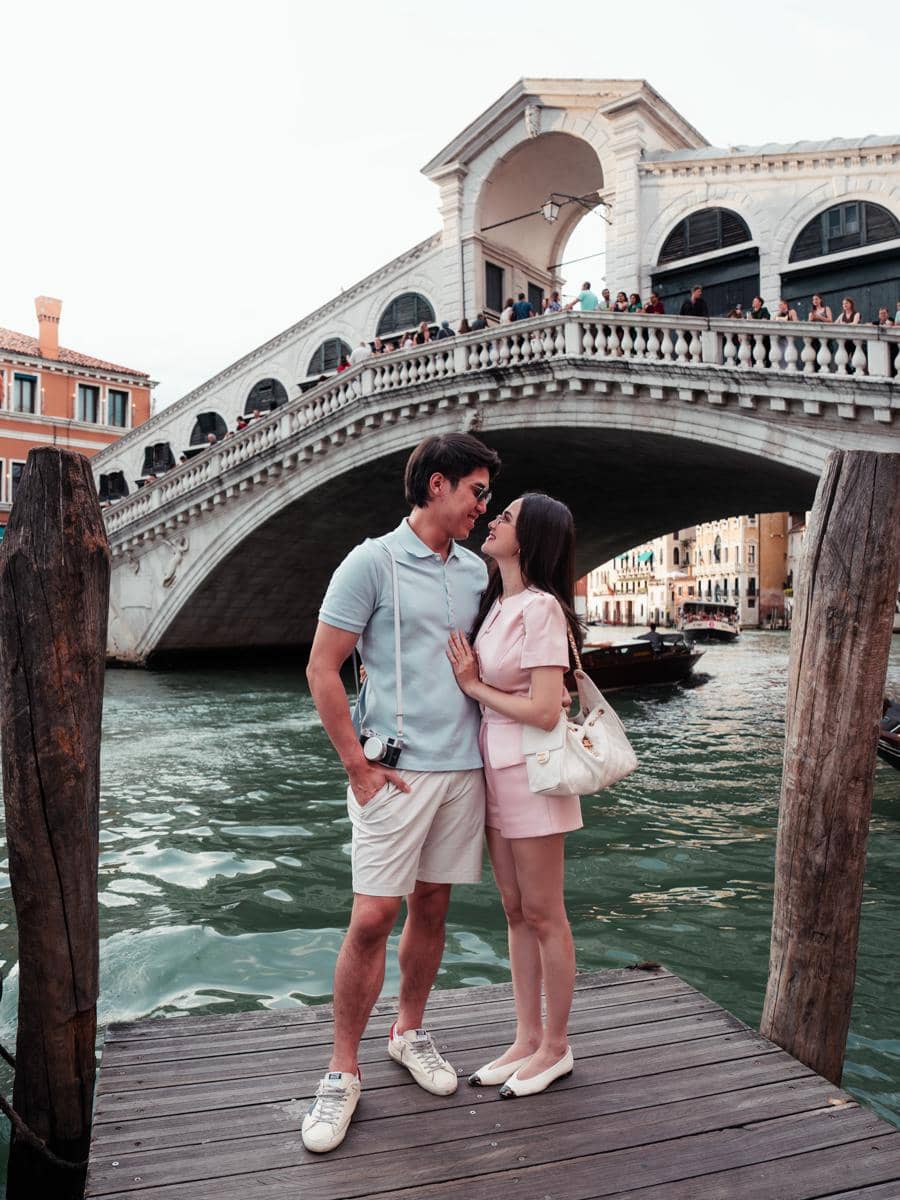 Sepasang kekasih berdiri di tepi kanal Venesia dengan latar Jembatan Rialto, saling menatap mesra di bawah langit cerah.