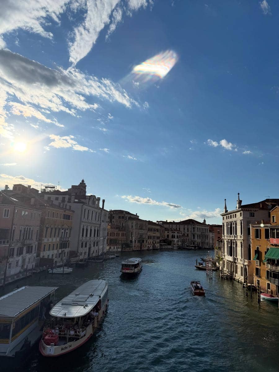 Pemandangan kanal di Venice dengan beberapa perahu melintas di bawah langit biru cerah dan bangunan klasik di tepi air.