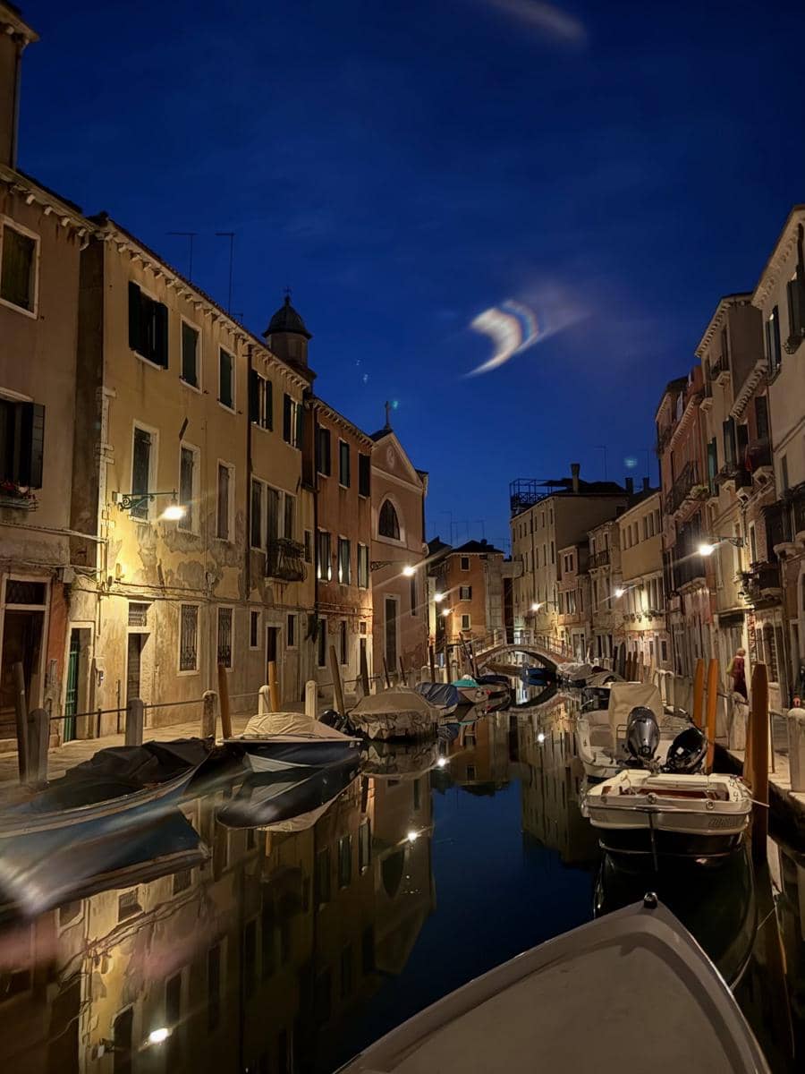 Pemandangan kanal di Venice pada malam hari dengan perahu yang terparkir di tepi bangunan berarsitektur klasik dan langit biru gelap.