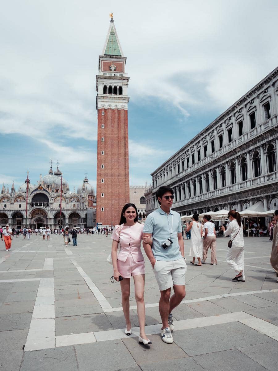 Sepasang wisatawan berjalan berpegangan tangan di Piazza San Marco, Venesia, dengan latar belakang Menara Campanile dan Basilika Santo Markus.