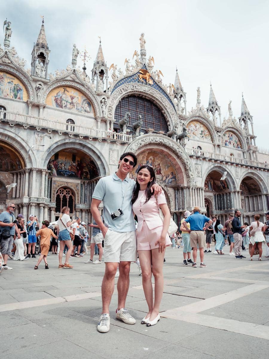 Sepasang wisatawan berpose di depan Basilika Santo Markus di Venesia, Italia, dengan suasana ramai turis di sekitar mereka.