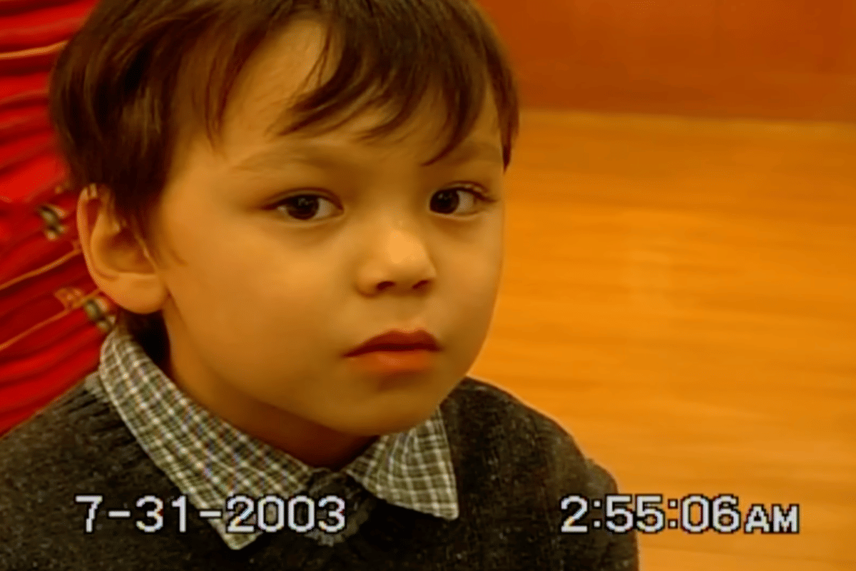 Seorang anak laki-laki mengenakan kemeja kotak-kotak dan sweater abu-abu, dengan latar belakang berwarna oranye hangat dan cap waktu 31 Juli 2003.