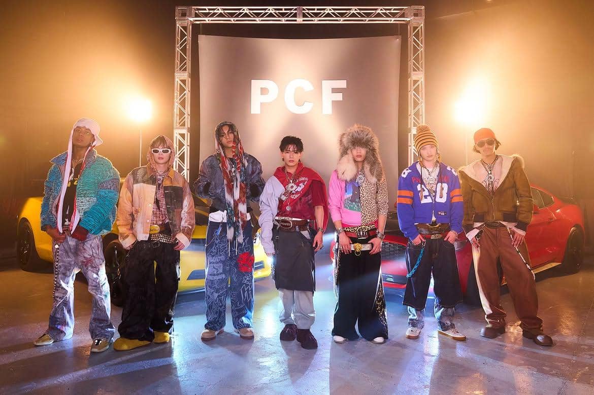 Tujuh personel boy band Jepang PSYCHIC FEVER berpose dengan busana bergaya streetwear di depan latar bertuliskan PCF dan mobil sport.