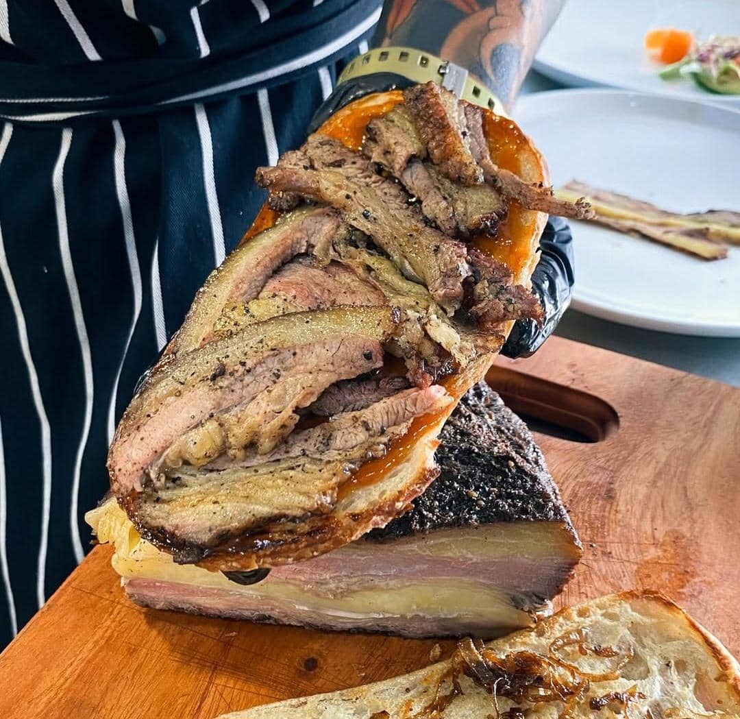 Potongan daging brisket asap tebal dengan lapisan lemak dan bumbu, dipegang oleh seseorang mengenakan sarung tangan hitam di atas talenan kayu.