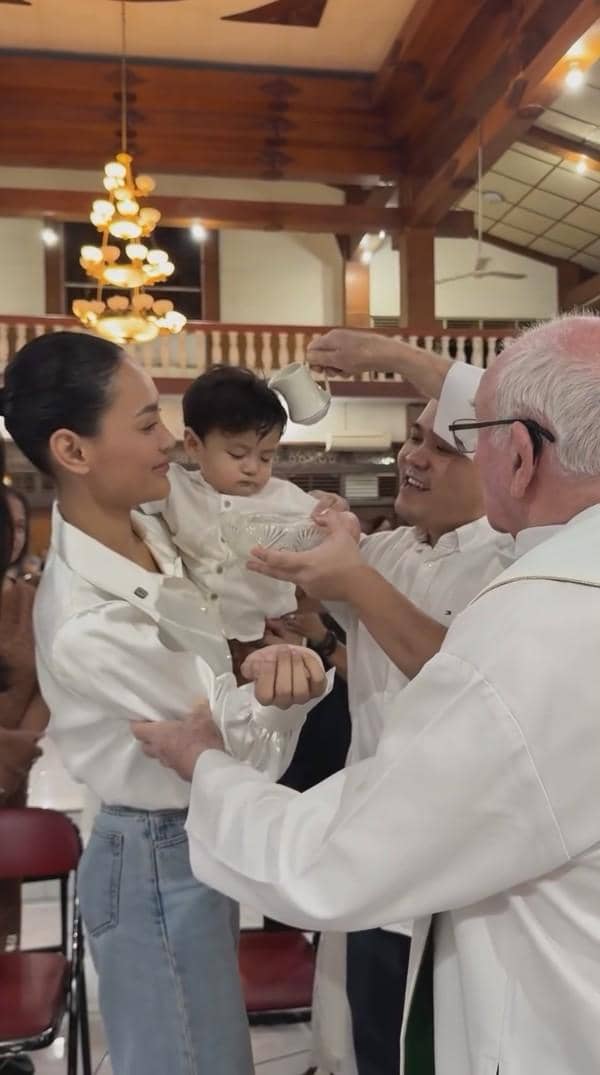 Seorang bayi sedang dibaptis di gereja, digendong oleh seorang wanita sambil seorang pendeta menuangkan air ke kepalanya.