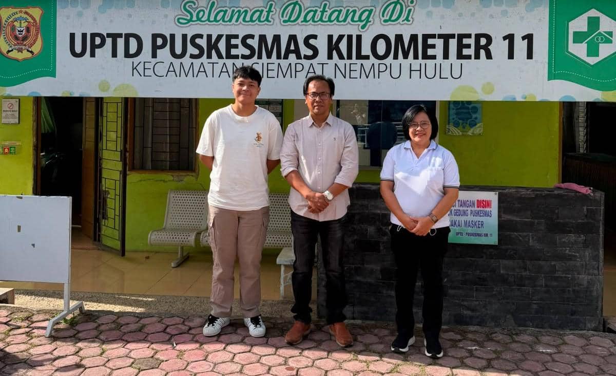 Tiga orang berdiri di depan papan nama UPTD Puskesmas Kilometer 11 Kecamatan Siempat Nempu Hulu, Kabupaten Dairi.