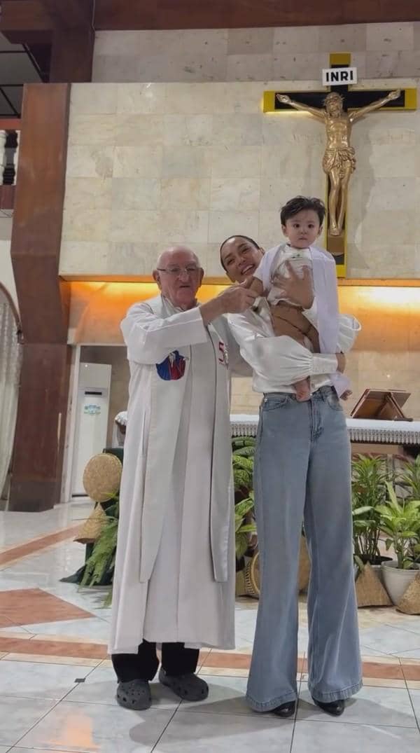 Seorang ibu bersama anak kecil dan seorang pastor berdiri di depan altar gereja dengan salib besar di latar belakang.