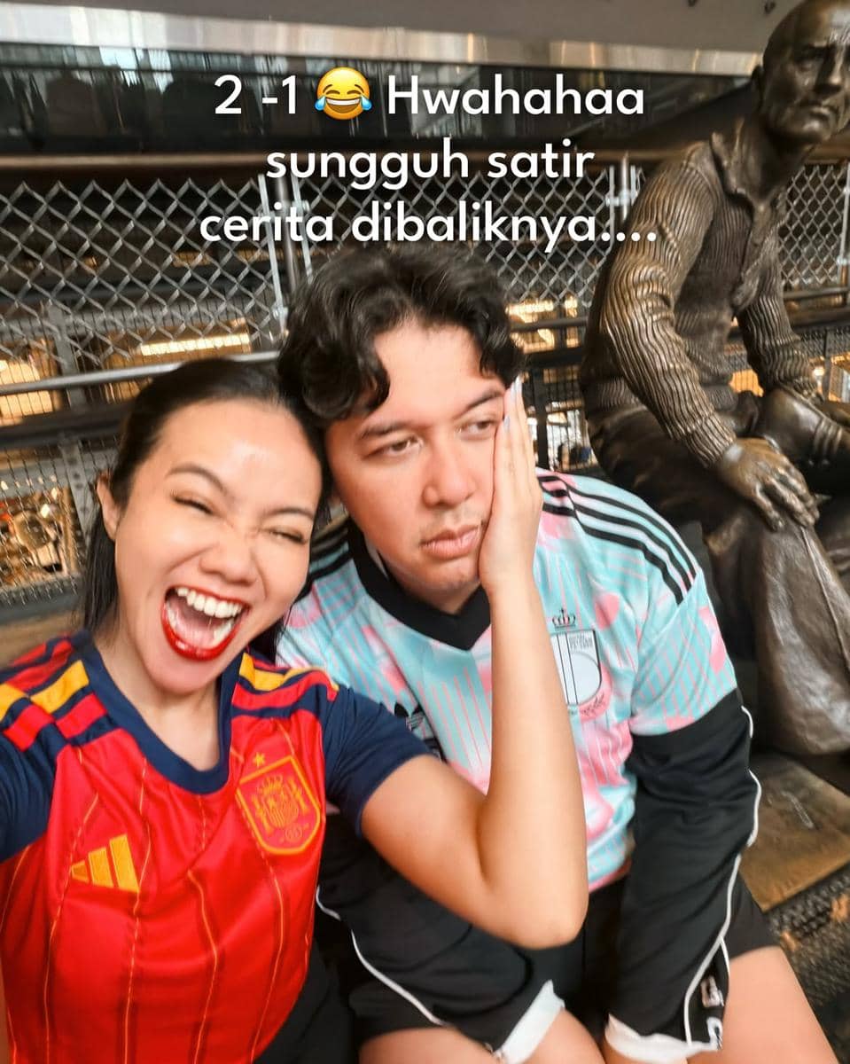 Seorang wanita mengenakan jersey tim Spanyol tertawa lebar sambil memegang wajah pria di sebelahnya yang mengenakan jersey tim lain dengan ekspresi datar.