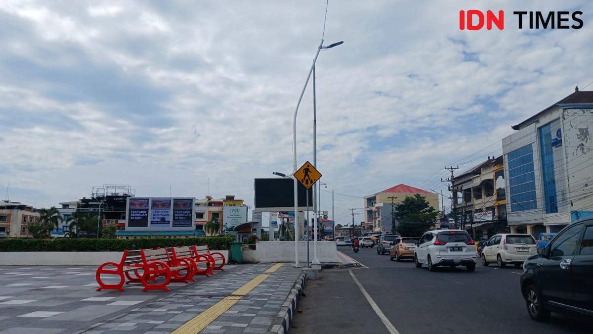 Lalu lintas kendaraan di jalan kota Manado dengan langit berawan dan deretan bangku merah di trotoar dekat area perkotaan.