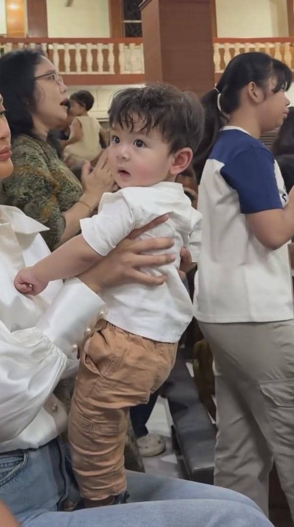 Seorang bayi mengenakan kemeja putih dan celana cokelat digendong oleh seorang wanita di dalam gereja saat acara baptisan.