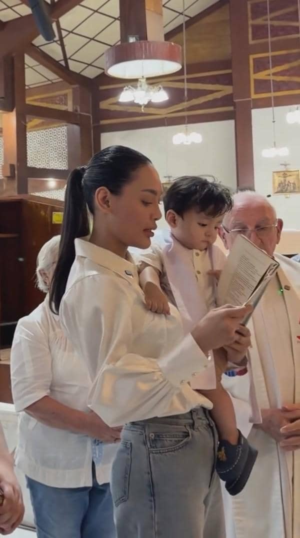 Seorang wanita mengenakan kemeja putih menggendong anak kecil saat prosesi pembaptisan di gereja bersama seorang pastor.