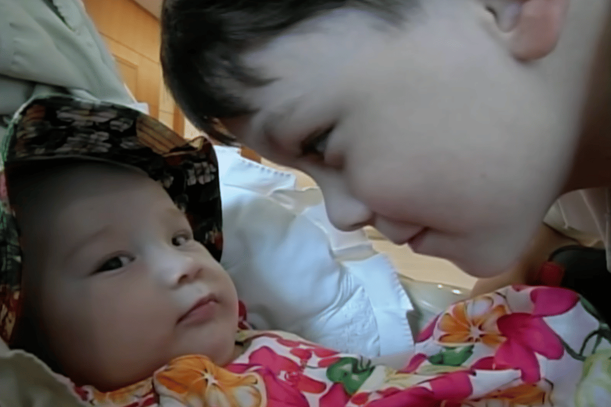 Seorang anak kecil menatap bayi yang mengenakan topi bermotif bunga dan pakaian berwarna cerah di dalam ruangan.