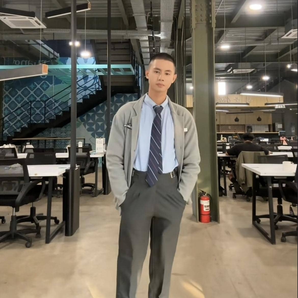 Seorang pria mengenakan kemeja biru, dasi, dan celana abu-abu berdiri di ruang kerja modern dengan meja dan kursi kantor di sekitarnya.