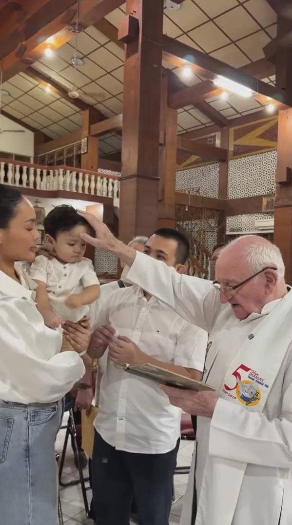 Seorang bayi mengenakan pakaian putih sedang dibaptis oleh seorang pastor di gereja, didampingi orang tua yang juga berpakaian putih.
