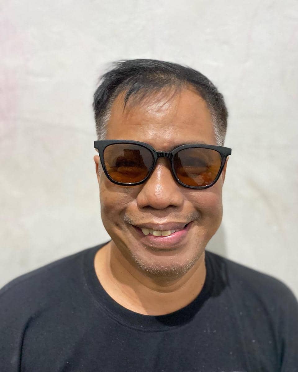 Seorang pria mengenakan kacamata hitam dan kaus hitam tersenyum di depan dinding berwarna terang dengan pencahayaan alami.