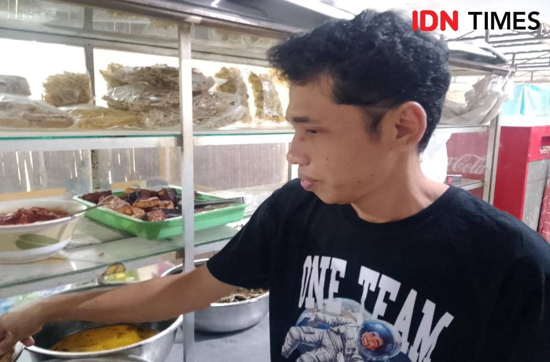 Seorang pria mengenakan kaus hitam sedang melayani pelanggan di warung makan dengan etalase berisi lauk pauk dan bahan makanan.